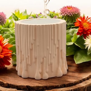May include: A white ceramic planter shaped like a tree trunk with a textured surface. The planter is surrounded by colorful flowers.