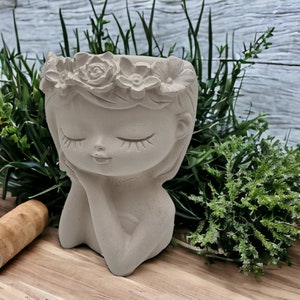May include: A gray concrete planter shaped like a girl's head with a flower crown. The planter has closed eyes and a serene expression.