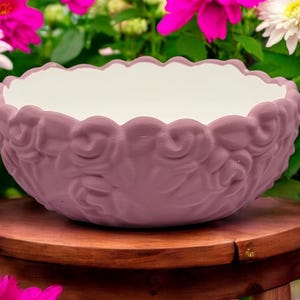 Könnte beinhalten: Ein mauvefarbener Keramiktopf mit weißem Innenraum und einem erhabenen Blumendesign. Der Topf steht auf einem Holzhocker.