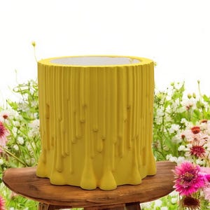 Könnte beinhalten: Ein gelber zylindrischer Pflanztopf mit einem strukturierten, tropfenden Design. Der Pflanztopf steht auf einer Holzoberfläche, umgeben von rosa und weißen Blumen. Der Pflanztopf ist etwa 30 cm hoch und 25 cm im Durchmesser.