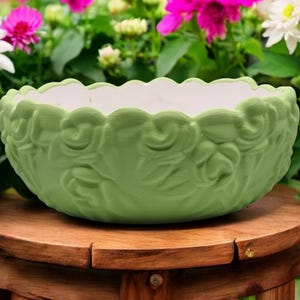 Op de afbeelding: Een groene keramische plantenbak met een bloemenpatroon. De plantenbak heeft een gegolfde rand en staat op een houten kruk.