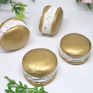 Op de afbeelding: Vier goudkleurige macarons met witte vulling. De macarons zijn in een halve cirkel op een wit oppervlak gerangschikt.