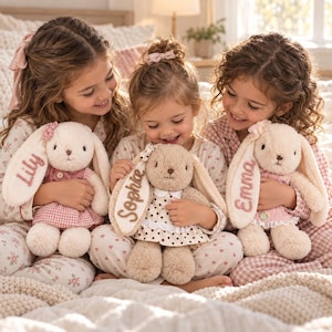 May include: Three plush stuffed rabbits with long ears, each wearing a dress and featuring an embroidered name on one ear. The left rabbit is cream with a pink dress, the center is tan with a polka dot dress, and the right is cream with a pink dress.