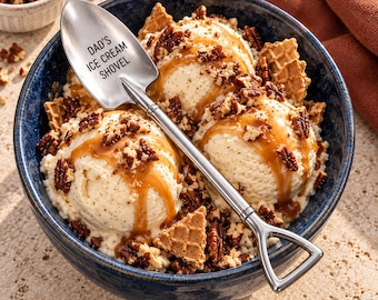 May include: A silver metal shovel-shaped spoon with the text DAD'S ICE CREAM SHOVEL engraved on the bowl. The spoon rests in a dark blue bowl filled with three scoops of vanilla ice cream, drizzled with caramel sauce, and topped with pecans.