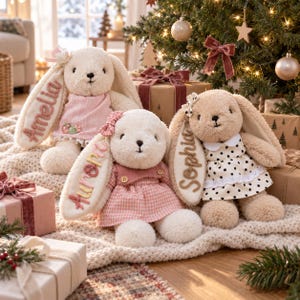 May include: Three plush cream-colored bunnies with long ears, each wearing a dress and featuring an embroidered name on one ear. The left bunny is named Amelia, the center bunny is Aurora, and the right bunny is named Sophia.