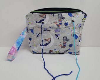 May include: A gray zippered tote bag with a mermaid and seahorse print. The bag has a long, colorful strap and two blue and turquoise yarn tails hanging from the top.