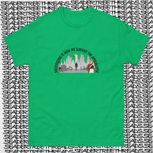 May include: A green t-shirt with a black graphic print of a cityscape with people walking. The text "Shitposting is how we survive the apocalypse" is printed in a semi-circle around the graphic.
