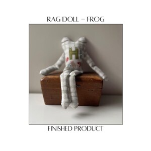 May include: A handmade rag doll frog with a gray and white checkered pattern. The frog has a green "H" on its chest and is sitting on a small wooden box. The text "RAG DOLL - FROG" and "FINISHED PRODUCT" are displayed.