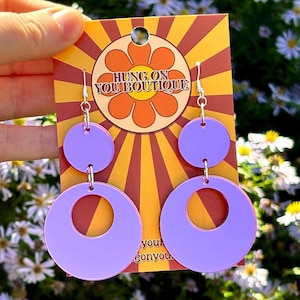 以下が含まれることがあります： 円形のデザインの、ライラック色のレトロスタイルのイヤリング。イヤリングは、「HUNG ON YOU BOUTIQUE」の文字と花のグラフィックが入ったカードから吊り下げられています。背景は花と葉の混合です。