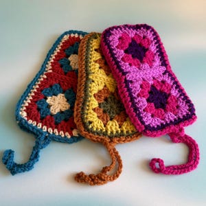 Peut inclure: Trois étuis de téléphone crochetés à la main et colorés. Les étuis présentent un motif de granny square dans les tons de bleu, rouge, jaune et rose. Chaque étui a une boucle en bas. Le fond est bleu clair.