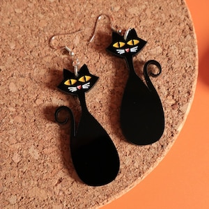 Pode incluir: Um par de brincos pretos em forma de gato. Cada brinco apresenta uma silhueta de gato estilizada com olhos amarelos, bigodes brancos e um pequeno nariz vermelho. Os brincos penduram-se em ganchos prateados.