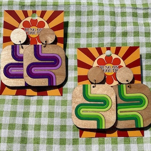 May include: Wooden square earrings with a retro design. One pair features purple and pink wavy lines, the other green and lime. Each earring has a small wooden circle at the top and is displayed on a card with the text "HUNG ON BOUTIQUE".