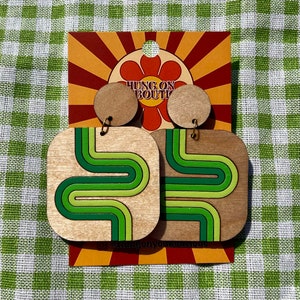 Puede incluir: Pendientes de madera con un dise&ntilde;o retro. Los pendientes tienen forma cuadrada con un patr&oacute;n de rayas verdes y verde claro onduladas. Los pendientes est&aacute;n sujetos a un perno redondo de madera. El texto "HUNG ON BOUTI" se muestra en una tarjeta.