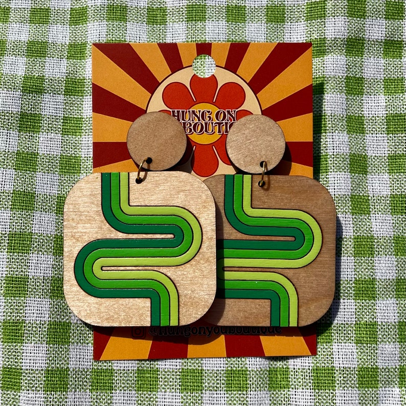 Puede incluir: Pendientes de madera con un dise&ntilde;o retro. Los pendientes tienen forma cuadrada con un patr&oacute;n de rayas verdes y verde claro onduladas. Los pendientes est&aacute;n sujetos a un perno redondo de madera. El texto "HUNG ON BOUTI" se muestra en una tarjeta.
