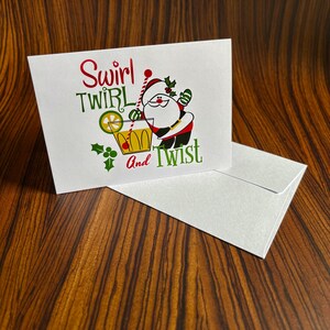 May include: A white Christmas card with a cartoon Santa Claus holding a glass of orange juice with a lime wedge. The text on the card reads "Swirl Twirl And Twist".