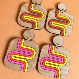 Puede incluir: Cuatro pares de pendientes de madera con un dise&ntilde;o retro. Cada pendiente presenta una forma cuadrada con una parte superior circular y un patr&oacute;n de rayas onduladas de colores en tonos marr&oacute;n, naranja, amarillo y rosa. Los pendientes se muestran sobre una superficie naranja.
