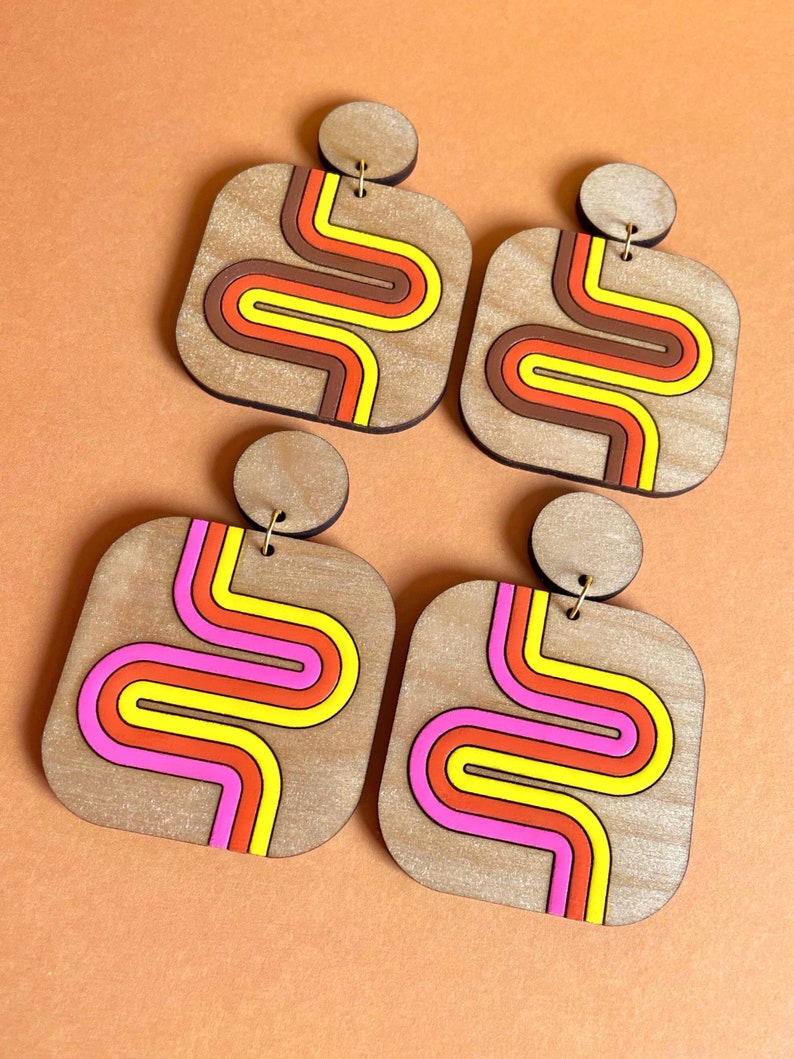 Puede incluir: Cuatro pares de pendientes de madera con un dise&ntilde;o retro. Cada pendiente presenta una forma cuadrada con una parte superior circular y un patr&oacute;n de rayas onduladas de colores en tonos marr&oacute;n, naranja, amarillo y rosa. Los pendientes se muestran sobre una superficie naranja.