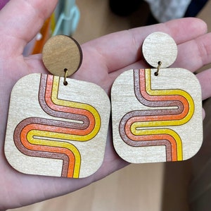 Puede incluir: Pendientes de madera con dise&ntilde;o retro. Cada pendiente tiene forma cuadrada con la parte superior redondeada y un perno circular. El dise&ntilde;o incluye un patr&oacute;n ondulado en naranja, amarillo y marr&oacute;n.