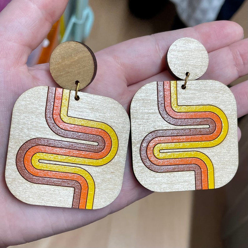 Puede incluir: Pendientes de madera con dise&ntilde;o retro. Cada pendiente tiene forma cuadrada con la parte superior redondeada y un perno circular. El dise&ntilde;o incluye un patr&oacute;n ondulado en naranja, amarillo y marr&oacute;n.