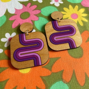 Puede incluir: Pendientes cuadrados de madera con dise&ntilde;o retro. Los pendientes presentan una veta de madera clara con un patr&oacute;n de ondas moradas, lavanda y magenta. Tienen un peque&ntilde;o perno redondo de madera en la parte superior y cuelgan de un alambre dorado.