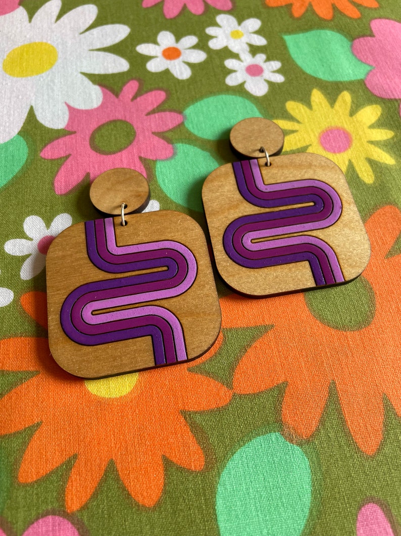 Puede incluir: Pendientes cuadrados de madera con dise&ntilde;o retro. Los pendientes presentan una veta de madera clara con un patr&oacute;n de ondas moradas, lavanda y magenta. Tienen un peque&ntilde;o perno redondo de madera en la parte superior y cuelgan de un alambre dorado.