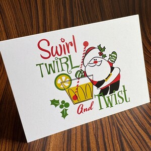 May include: A Christmas card with a cartoon Santa Claus holding a glass of a red and yellow drink with a lime wedge. The text on the card reads "Swirl Twirl and Twist".
