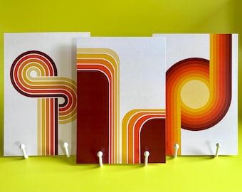 Puede incluir: Tres impresiones artísticas de estilo retro con diseños de rayas curvas y coloridas en tonos naranja, amarillo y marrón sobre un fondo blanco. Cada impresión tiene un gancho blanco en la parte inferior. Las impresiones se muestran sobre un fondo amarillo-verdoso brillante.
