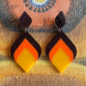 Peut inclure: Une paire de boucles d'oreilles pendantes de style rétro. Elles présentent un design superposé avec un dessus marron foncé, suivi de sections orange et jaune. Les boucles sont suspendues à de petits crochets dorés.