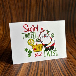 May include: A white card with a cartoon Santa Claus holding a glass of yellow liquid with a lime wedge and a red cherry. The text on the card reads "Swirl Twirl And Twist".