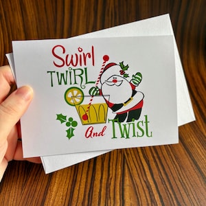 May include: A white Christmas card with a cartoon Santa Claus holding a cocktail glass. The text on the card reads "Swirl Twirl and Twist".