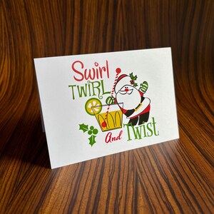 May include: A white card with a red and green illustration of Santa Claus holding a glass of a yellow drink with a lime wedge. The text on the card reads "Swirl Twirl and Twist".