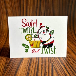 May include: A white card with a cartoon Santa Claus holding a cocktail glass with a lime wedge and a red cherry. The text on the card reads "Swirl Twirl And Twist".