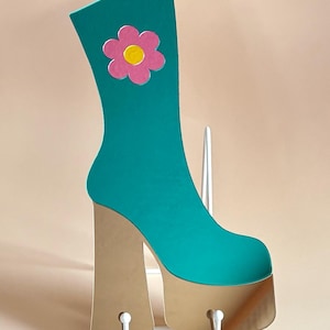 Peut inclure: Présentoir en forme de botte à plateforme turquoise avec une fleur rose et jaune. La botte a une plateforme et un talon beiges, soutenus par une structure métallique blanche. Le design rappelle la mode des années 1970.