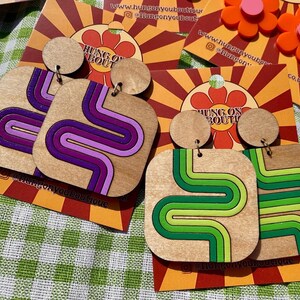 Puede incluir: Pendientes de madera con dise&ntilde;o retro. Un par presenta un patr&oacute;n ondulado morado y rosa, y el otro tiene un patr&oacute;n ondulado verde y verde lima. Cada pendiente tiene una parte superior redonda peque&ntilde;a y una parte inferior cuadrada m&aacute;s grande. Los pendientes est&aacute;n sobre un fondo estampado.