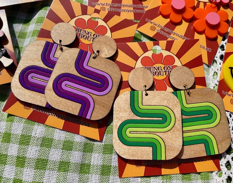 Puede incluir: Pendientes de madera con dise&ntilde;o retro. Un par presenta un patr&oacute;n ondulado morado y rosa, y el otro tiene un patr&oacute;n ondulado verde y verde lima. Cada pendiente tiene una parte superior redonda peque&ntilde;a y una parte inferior cuadrada m&aacute;s grande. Los pendientes est&aacute;n sobre un fondo estampado.