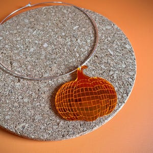 May include: A silver choker necklace with an orange, mirrored pumpkin charm. The pumpkin is a geometric design with a grid pattern.