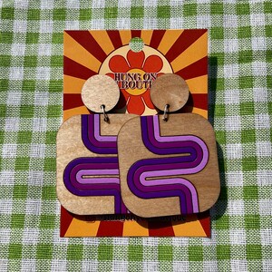 Puede incluir: Pendientes de madera con dise&ntilde;o retro. Los pendientes tienen forma cuadrada con un patr&oacute;n ondulado en tonos morados. Los pendientes est&aacute;n sujetos a un perno de madera redondo. El fondo es una tarjeta de estilo retro con el texto "HUNG ON ABOUT".