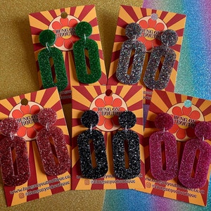 May include: Four pairs of dangle earrings with glitter and a rectangular shape. The earrings are displayed on cards with the text "Hung On Boutique" and a daisy design. The earrings are in the colors green, silver, black, and pink.