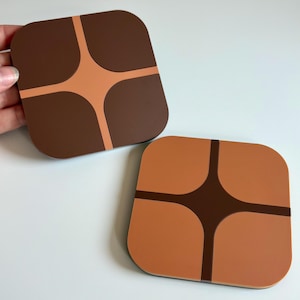 May include: Two square coasters with rounded corners. One is brown with a light brown cross design, and the other is light brown with a dark brown cross design. The coasters are on a white surface.