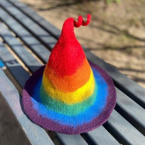 Pode incluir: Um chapéu de bruxa arco-íris de feltro. O chapéu tem uma aba larga e uma coroa cónica, com faixas vermelhas, laranja, amarelas, verdes, azuis e roxas. Uma espiral vermelha adorna o topo.