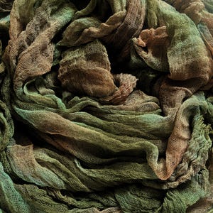 May include: Close-up of a textured fabric with a layered, draped appearance. The fabric features a blend of earthy tones, including shades of brown, green, and hints of teal. The material appears to be lightweight and airy, with visible woven details.