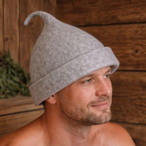 May include: A light grey sauna hat with a curved, pointed top and a rolled brim. The hat is made of a soft, felt-like material. The background is a wooden wall.