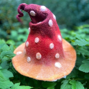 May include: A whimsical, handmade mushroom hat. The hat features a vibrant red cap with white polka dots, transitioning to an orange brim. The hat's design includes a curled tip, adding to its playful aesthetic. The hat is set against a backdrop of green foliage.