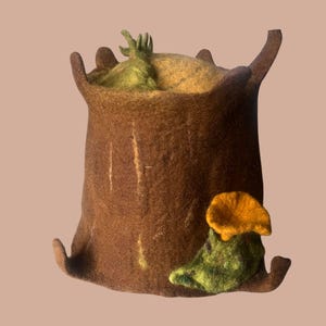 Könnte beinhalten: Ein handgefertigter Filzhut in Form eines Baumstumpfes, mit brauner Außenseite und gelb-grüner Innenseite. Ein Filzpilz mit orangefarbenem Hut und grünem Stiel ist seitlich angebracht. Der Hut hat ein skurriles, von der Natur inspiriertes Design.