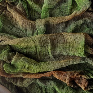 May include: Close-up of a textured fabric with a crinkled appearance. The fabric features a gradient of colors, transitioning from shades of green to brown. The material appears to be lightweight and draped, with visible folds and shadows.