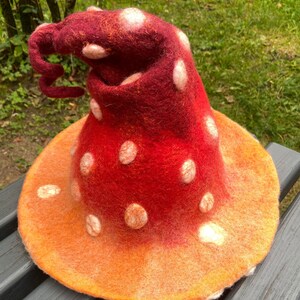 May include: A whimsical, handmade felt hat in the shape of a mushroom. The hat features a vibrant red cap with white spots and an orange brim. The cap has a curled tip, adding to its unique design. This hat is perfect for costume parties or themed events.
