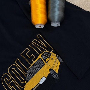 May include: A black t-shirt featuring an embroidered yellow car and the text GOLF IV in yellow thread, accompanied by two spools of thread in matching yellow and dark gray colors placed at the top of the garment fabric.
