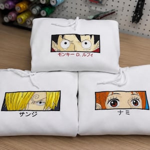 May include: Three white hooded sweatshirts featuring embroidered rectangular patches of anime character eyes. The top hoodie shows Monkey D. Luffy, the bottom left shows Sanji, and the bottom right shows Nami, each with their name in Japanese text.