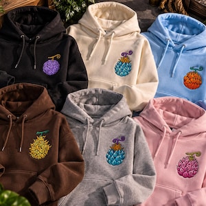 Puede incluir: Una colección de seis sudaderas con capucha en negro, crema, azul claro, marrón, gris y rosa. Cada sudadera cuenta con un bolsillo tipo canguro frontal, cordones a juego y un gráfico bordado de una fruta estilizada con un patrón de espiral.