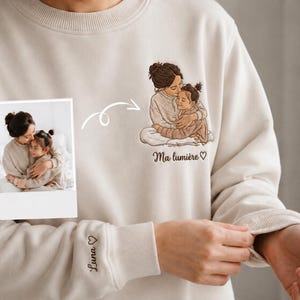 May include: A beige crewneck sweatshirt featuring an embroidered illustration of an adult and a child embracing. The text "Ma lumière" with a heart is embroidered below the design, and the name "Luna" with a heart is on the sleeve cuff.